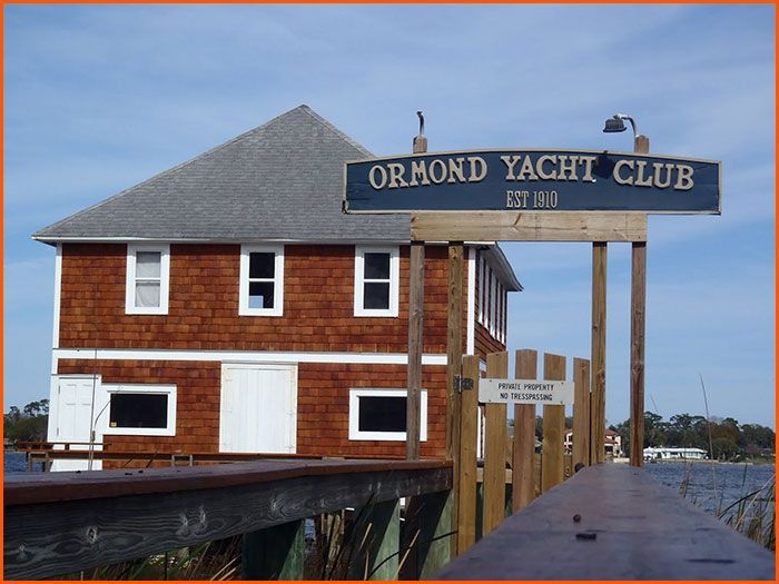 Ormond Yacht Club building on a pier with sign, brown cedar siding, blue sky, and water.