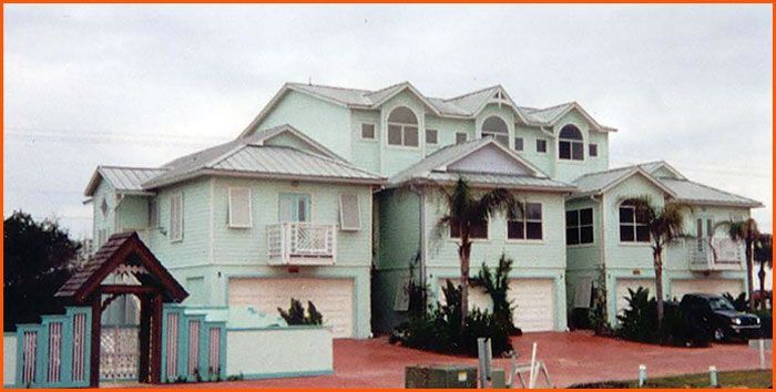 Multi-story teal beach house with gray roofs and red brick driveway.