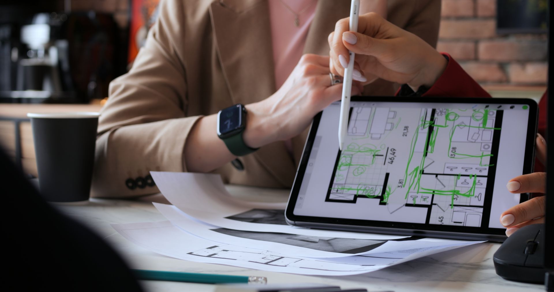 People reviewing a floor plan on a tablet, using a stylus. Papers and a coffee cup on the table.