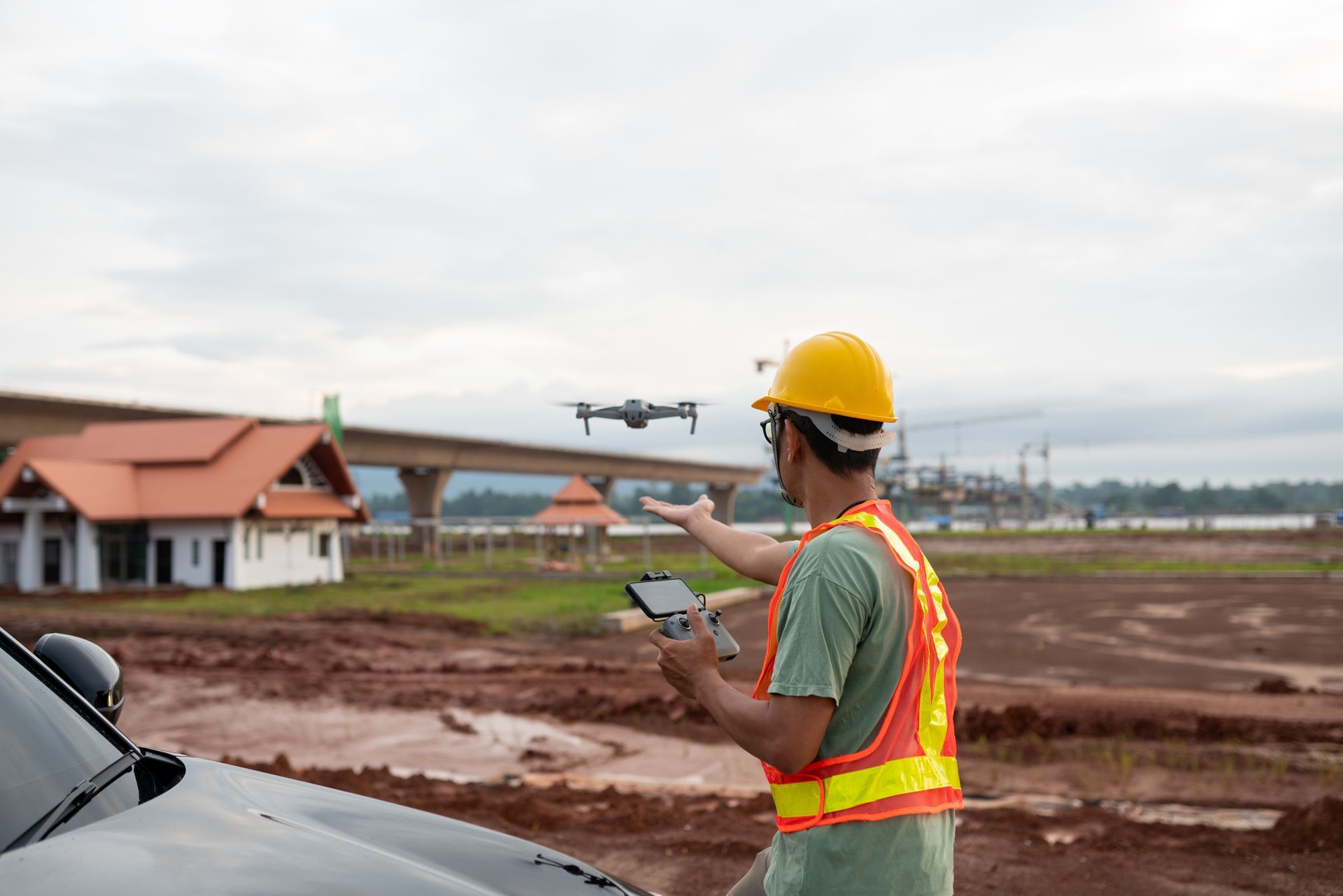 Construction worker operating drone at a construction site. Yellow hard hat, safety vest. Drone in air, overcast sky.