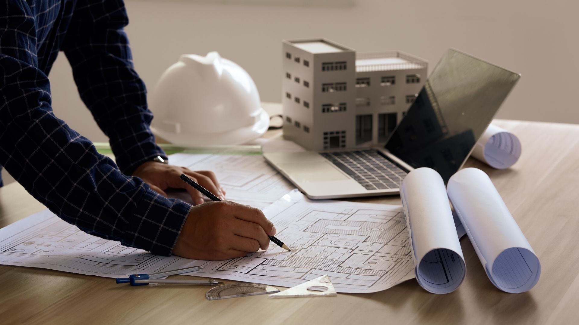 Architect working on blueprints at a desk with a model building, laptop, rolled plans, and a hard hat.