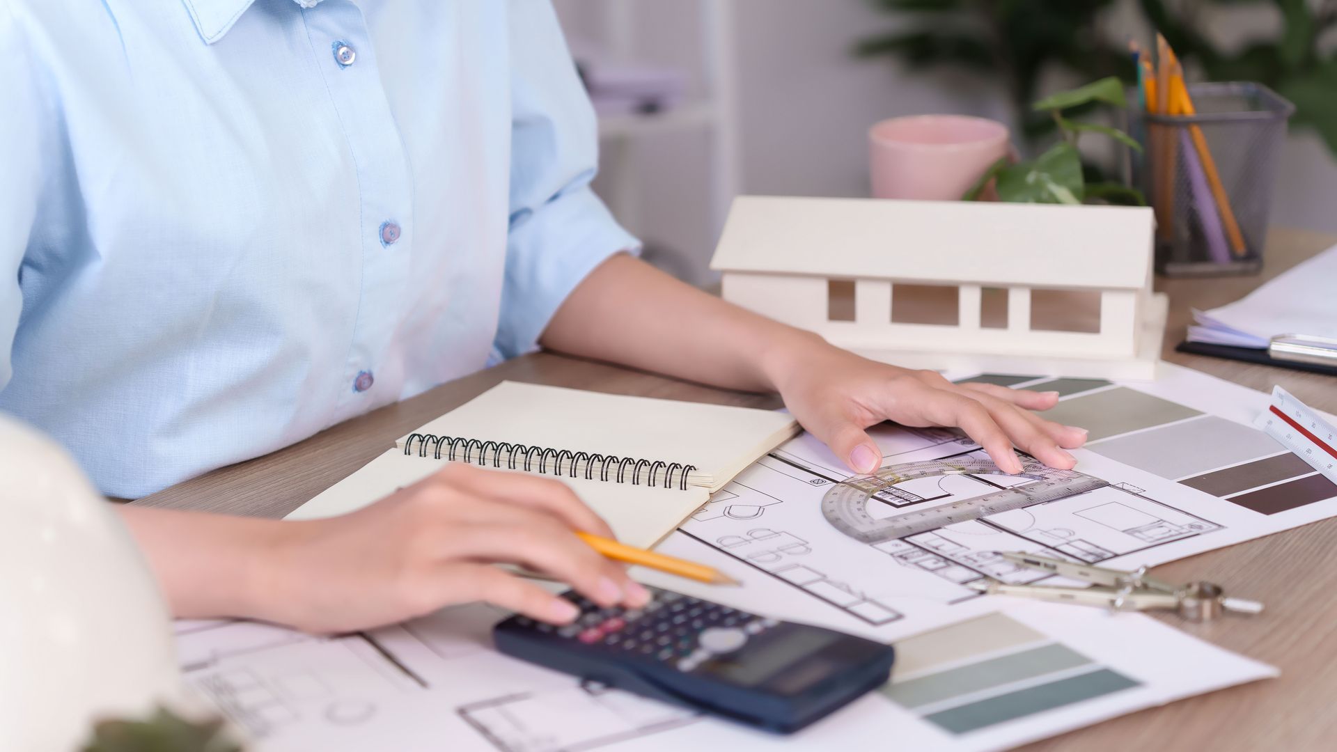 Architect working on blueprints, calculating with a calculator, with a model house and color swatches on a desk.