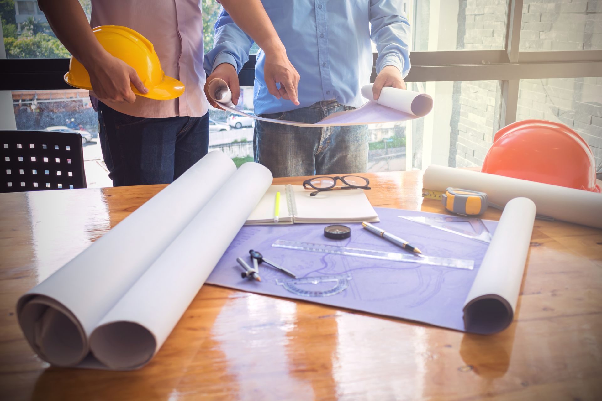 Two people reviewing blueprints at a table, hard hats, drawing tools, and rolled plans.