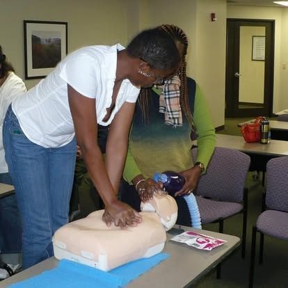 Woman demonstrating CPR on a mannequin, another woman assisting, in a classroom setting.