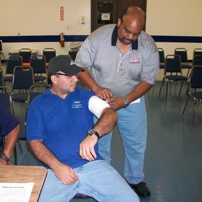 Man in blue shirt has arm examined by another man in a grey shirt indoors at a training.