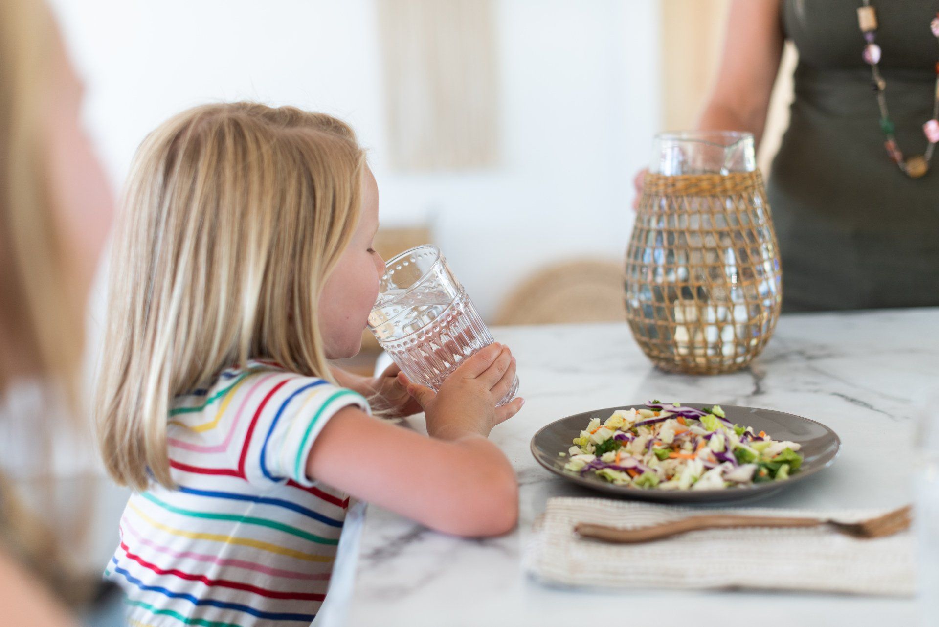 A little girl is drinking water from a glass while sitting at a table.