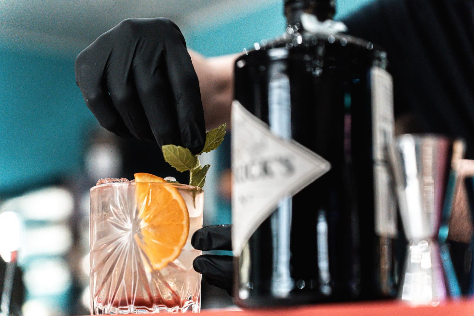 Un barista sta preparando un drink con una bottiglia di gin sullo sfondo.