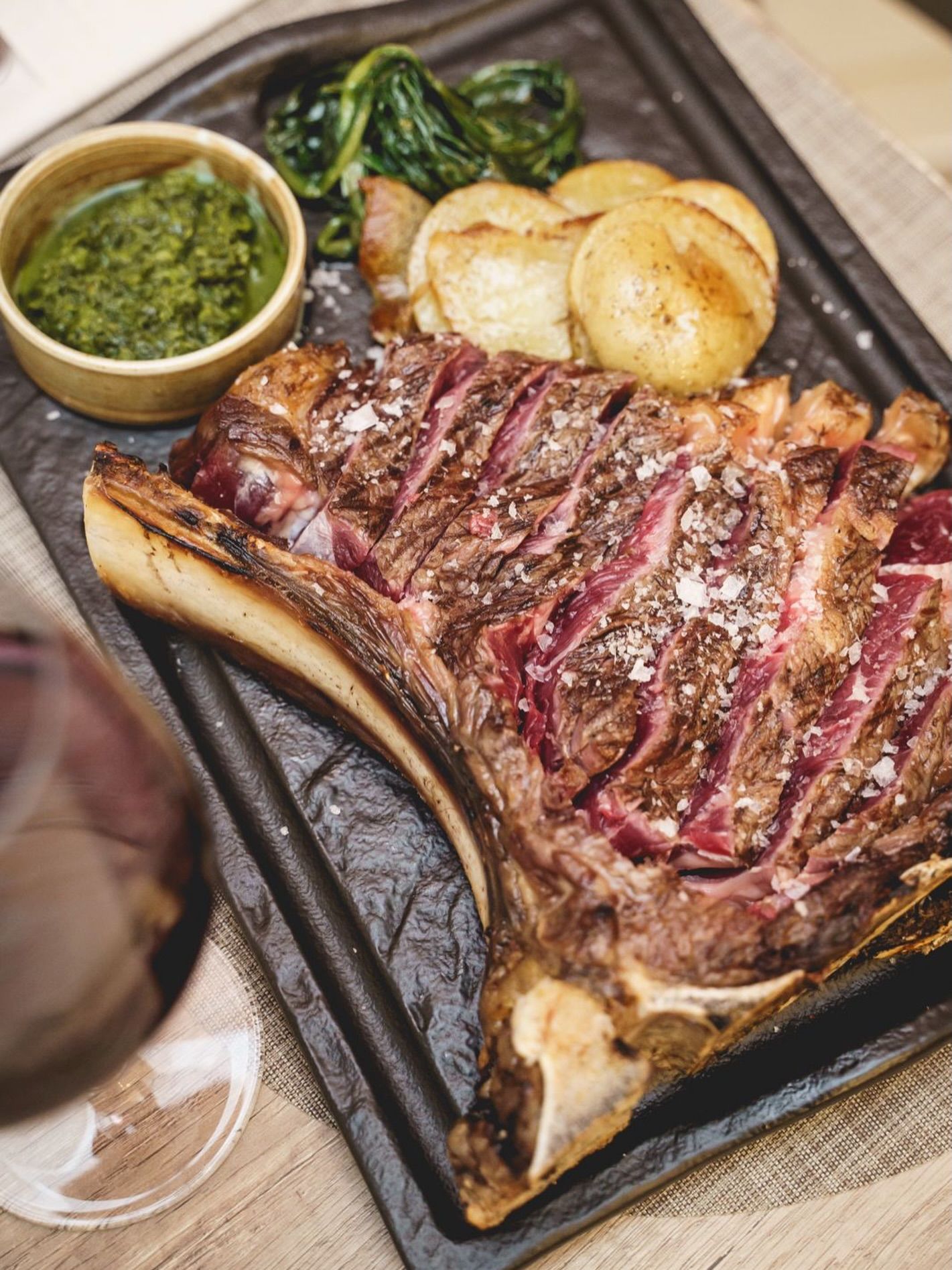 Bistecca tomahawk alla griglia con patate, verdure e chimichurri su un piatto nero.