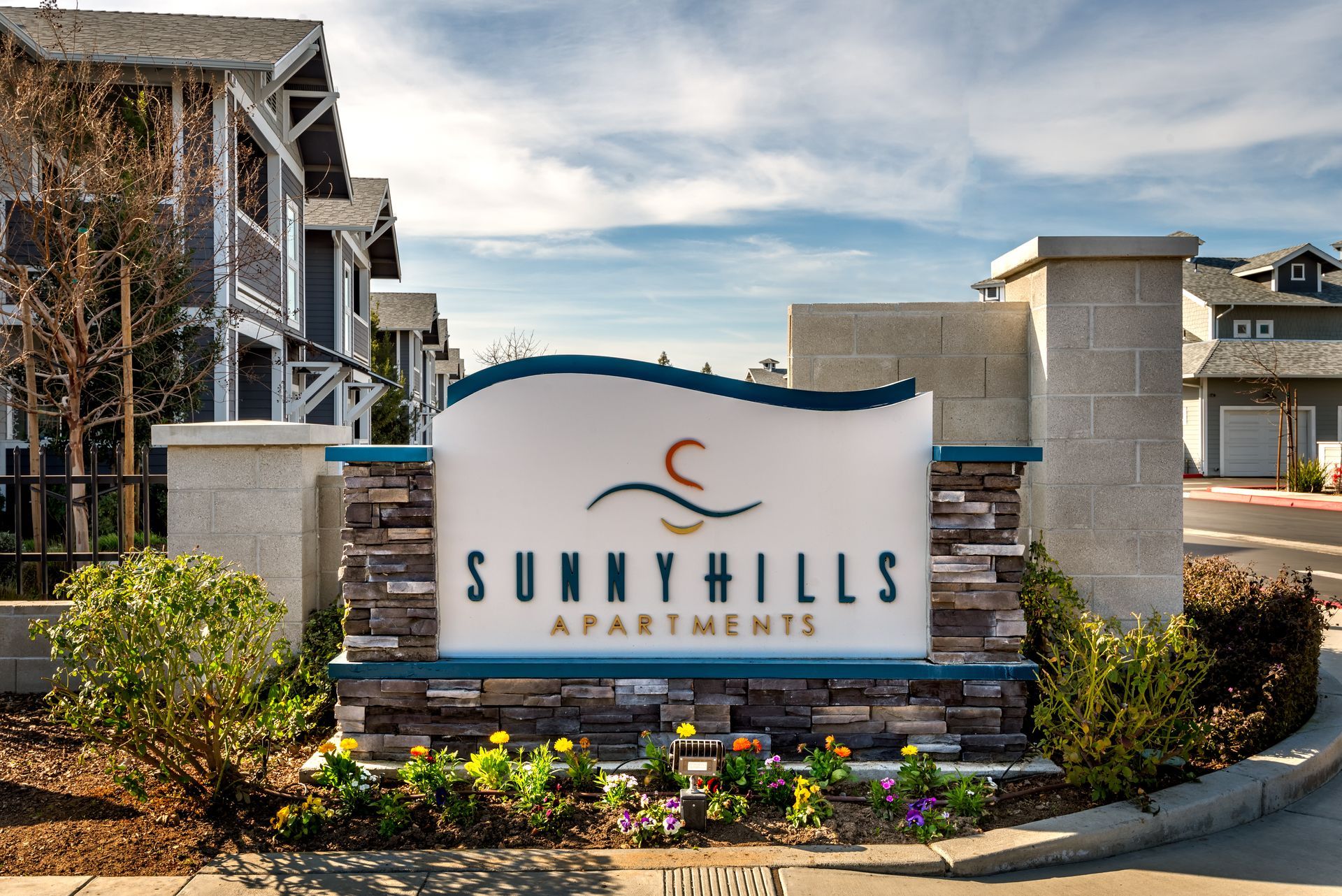 Sunny Hills Apartments entrance sign with blue accents, stone columns, and floral landscaping.