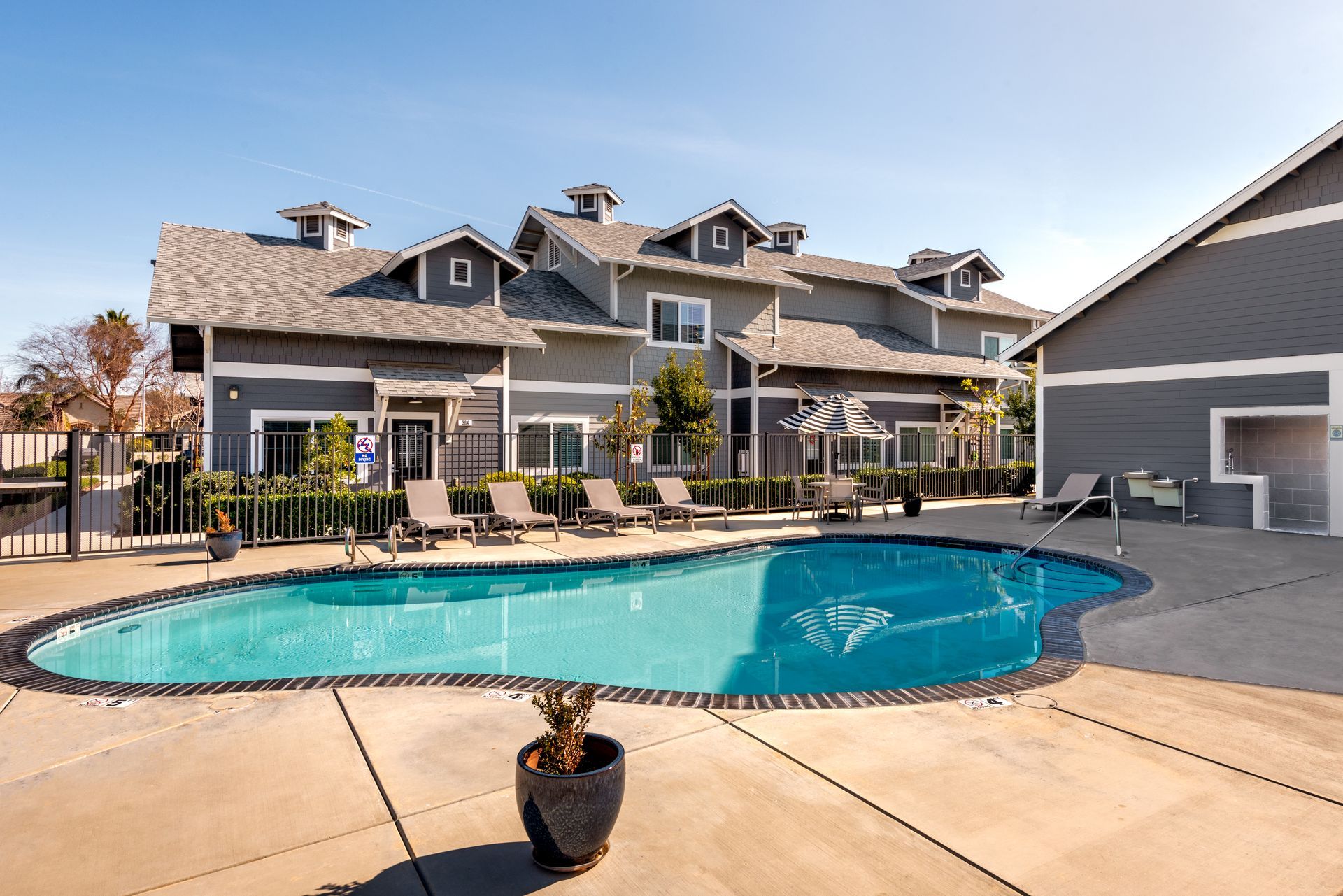 Swimming pool in front of gray multi-unit building with lounge chairs. Sunny day.