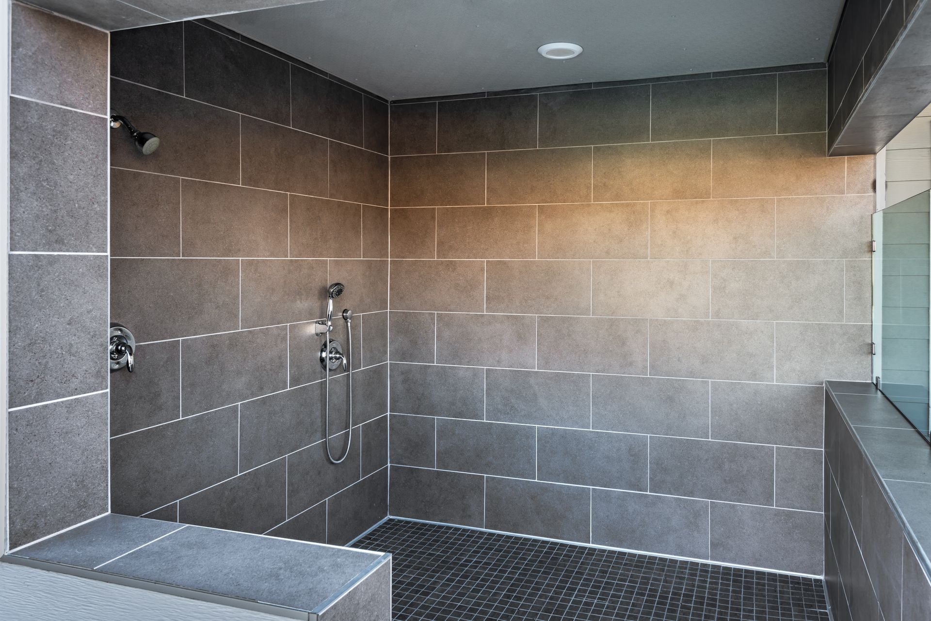 Dark tiled shower with a bench, glass wall, and pebble floor.