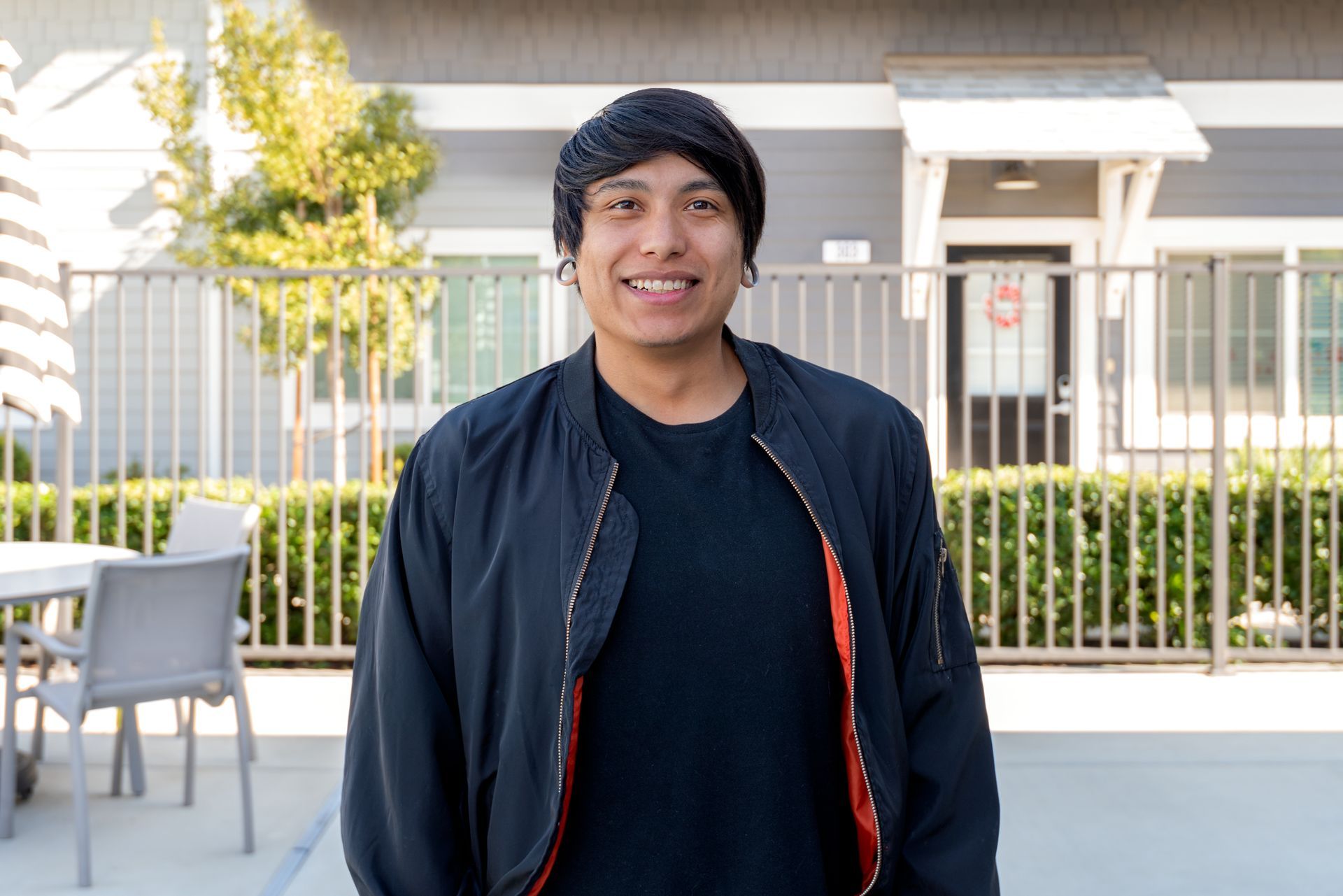 Young person with black hair, smiling, wearing a black jacket and dark shirt, standing outside by a fence.