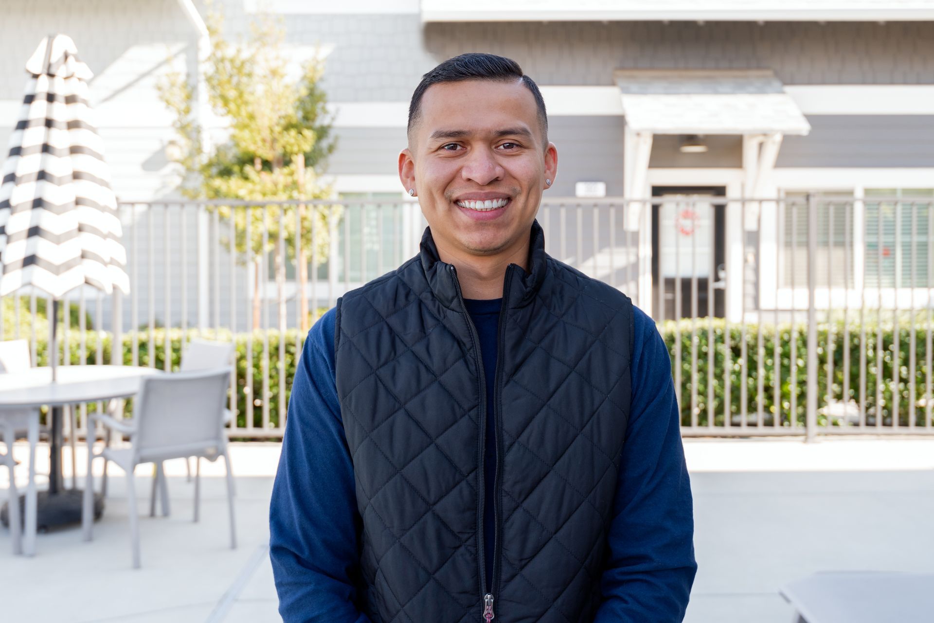 Man in a black vest smiles outdoors, in front of an apartment building.
