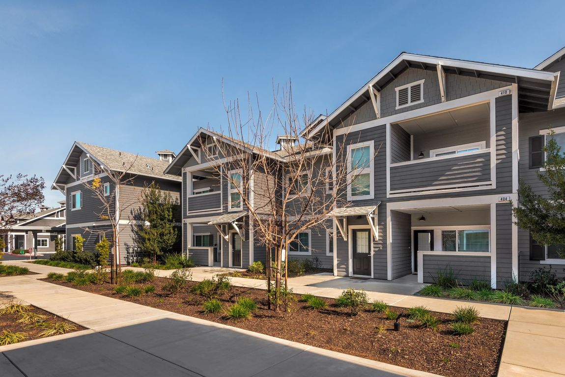 Gray two-story townhouses with balconies, brown landscaping, and a sidewalk on a sunny day.