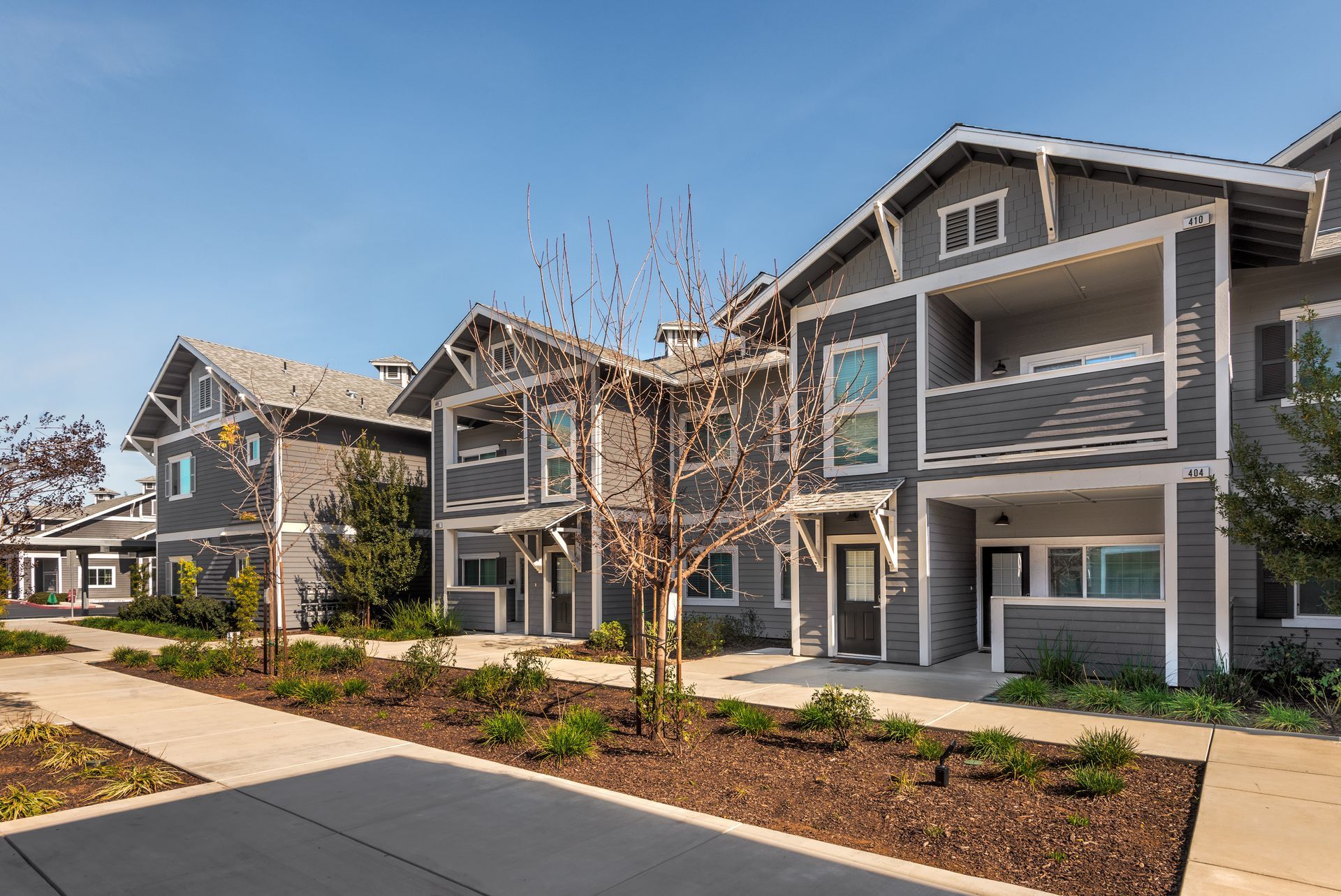 Gray two-story townhouses with balconies, brown landscaping, and a sidewalk on a sunny day.
