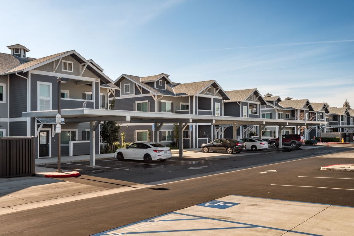 Row of gray apartment buildings with carports and parked cars under a blue sky.