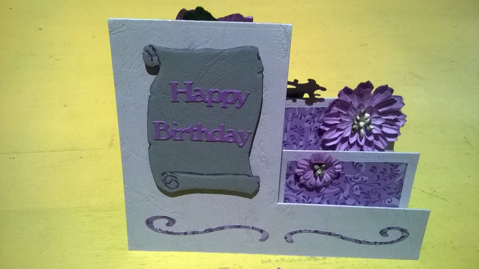 Eine Geburtstagskarte mit lila Blumen und einer Schriftrolle mit der Aufschrift „Happy Birthday“