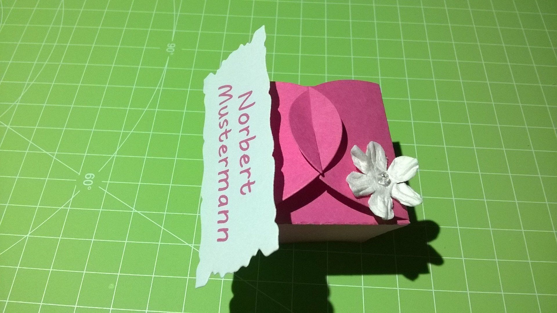 Eine rosa Schachtel mit einer Blume darauf steht auf einem grünen Schneidebrett.