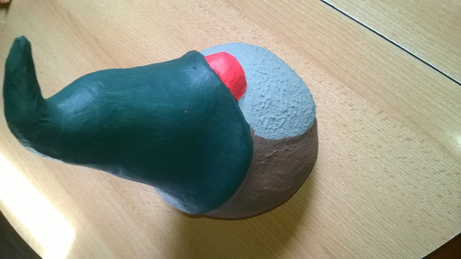 Auf einem Holztisch liegt ein grüner Hut mit einer roten Nase.