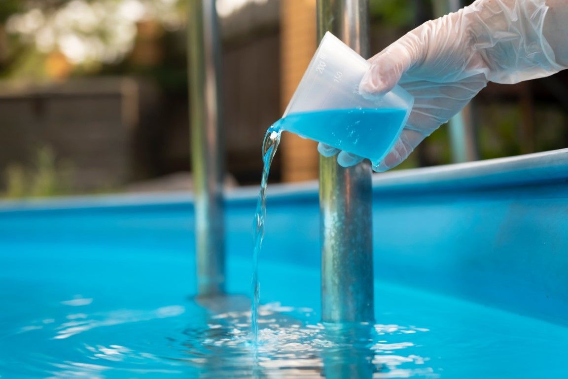 Hand pouring blue pool water test solution into a swimming pool beside a metal ladder