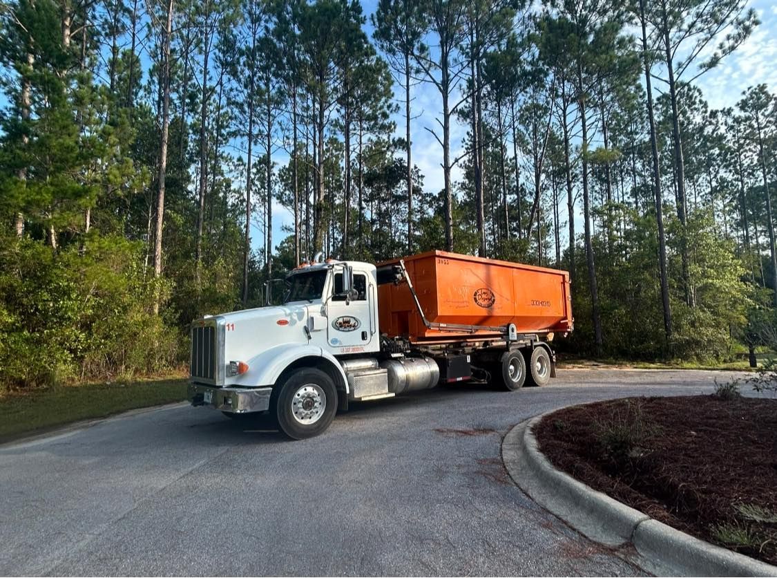 White dump truck with orange trailer driving on a curved road beside tall pine trees. White dump truck with orange trailer driving on a curved road beside tall pine trees.