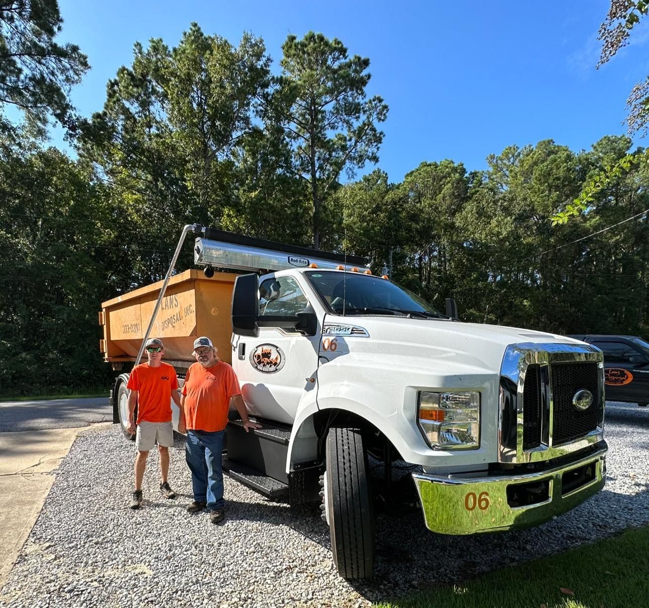 Two people in orange shirts standing beside a white dump truck on a sunny gravel road, surrounded by trees Two people in orange shirts standing beside a white dump truck on a sunny gravel road, surrounded by trees