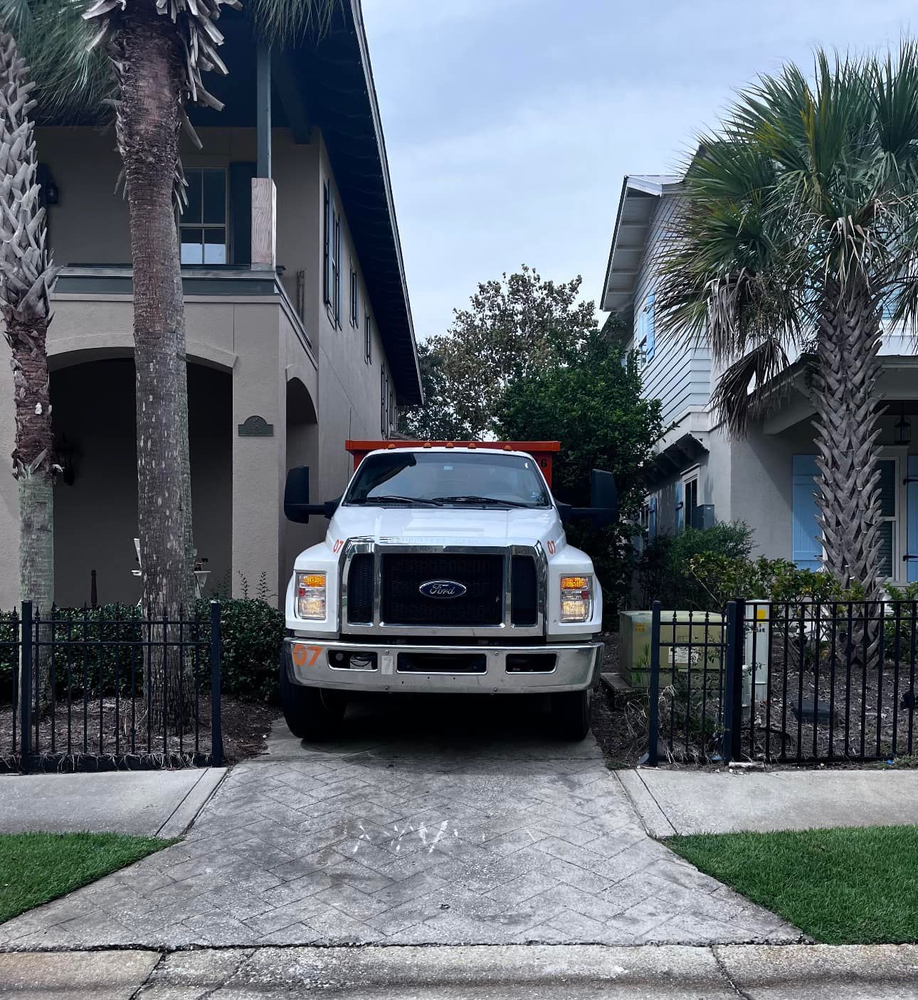 White Ford truck parked in a driveway between two houses, with palm trees and a black fence.