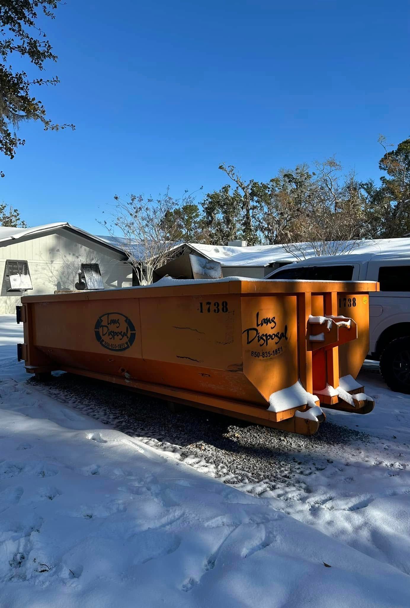 Orange dumpster in a snowy yard beside a house under a clear blue sky.