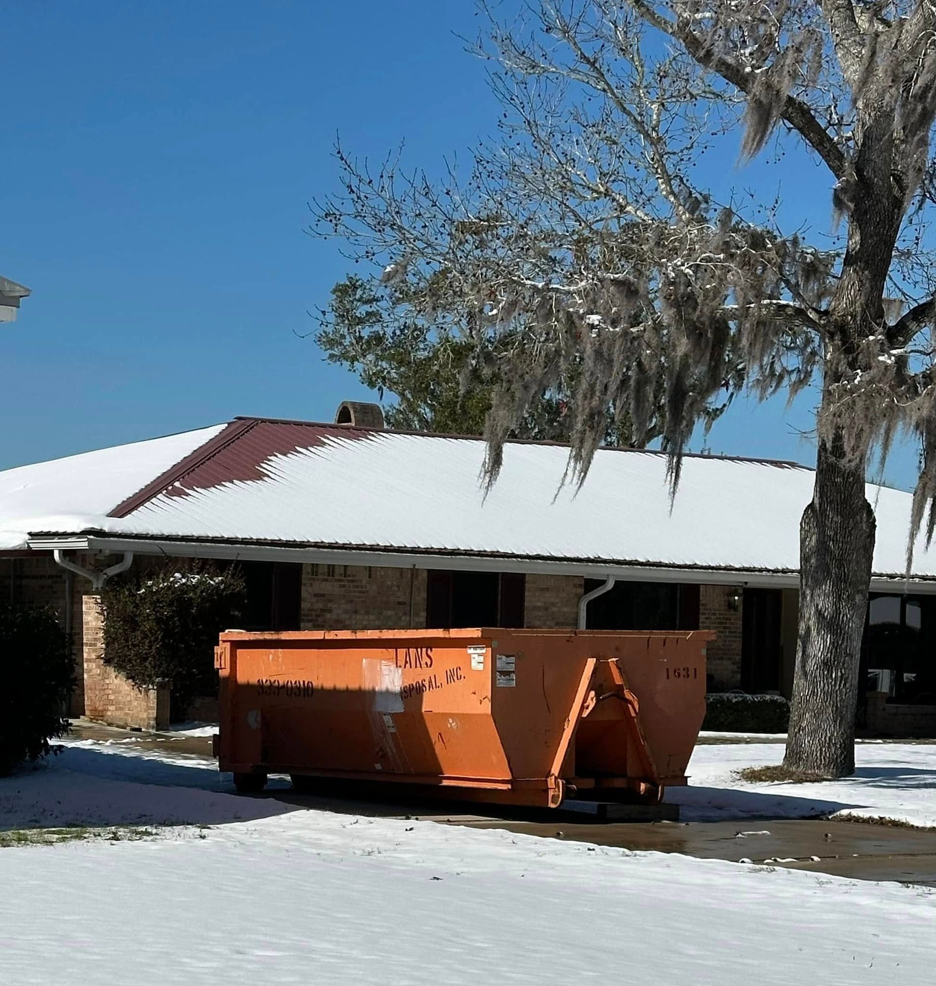 Snowy driveway with an orange dumpster in front of a house with a white roof and bare trees
