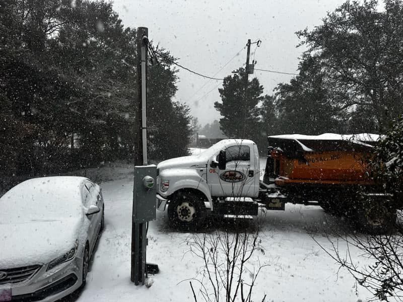 Snow-covered truck and car parked outside in a snowy driveway with trees and a utility pole. Snow-covered truck and car parked outside in a snowy driveway with trees and a utility pole.