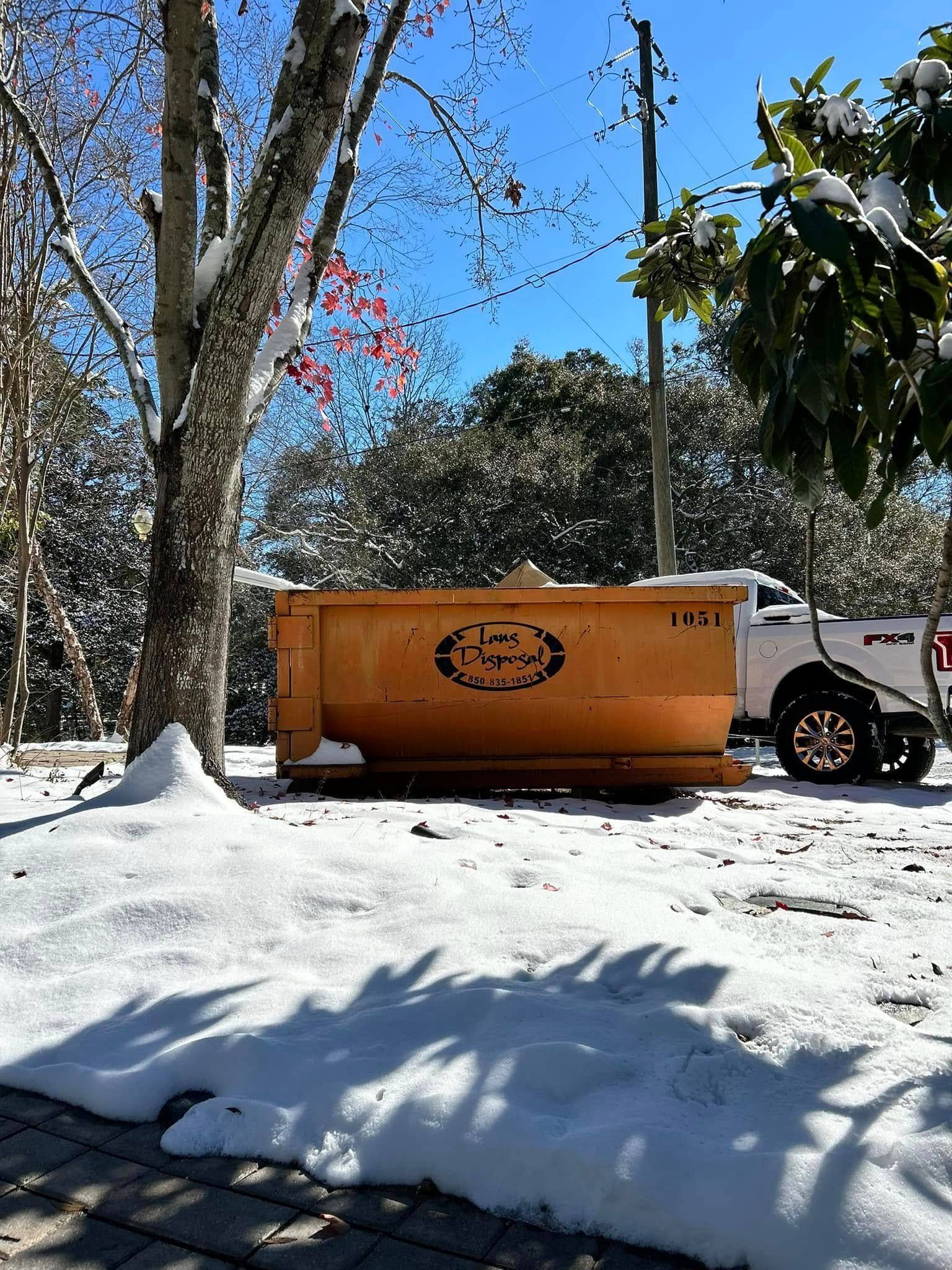 Orange dumpster in snowy yard beside trees and a white pickup truck under a blue sky Orange dumpster in snowy yard beside trees and a white pickup truck under a blue sky