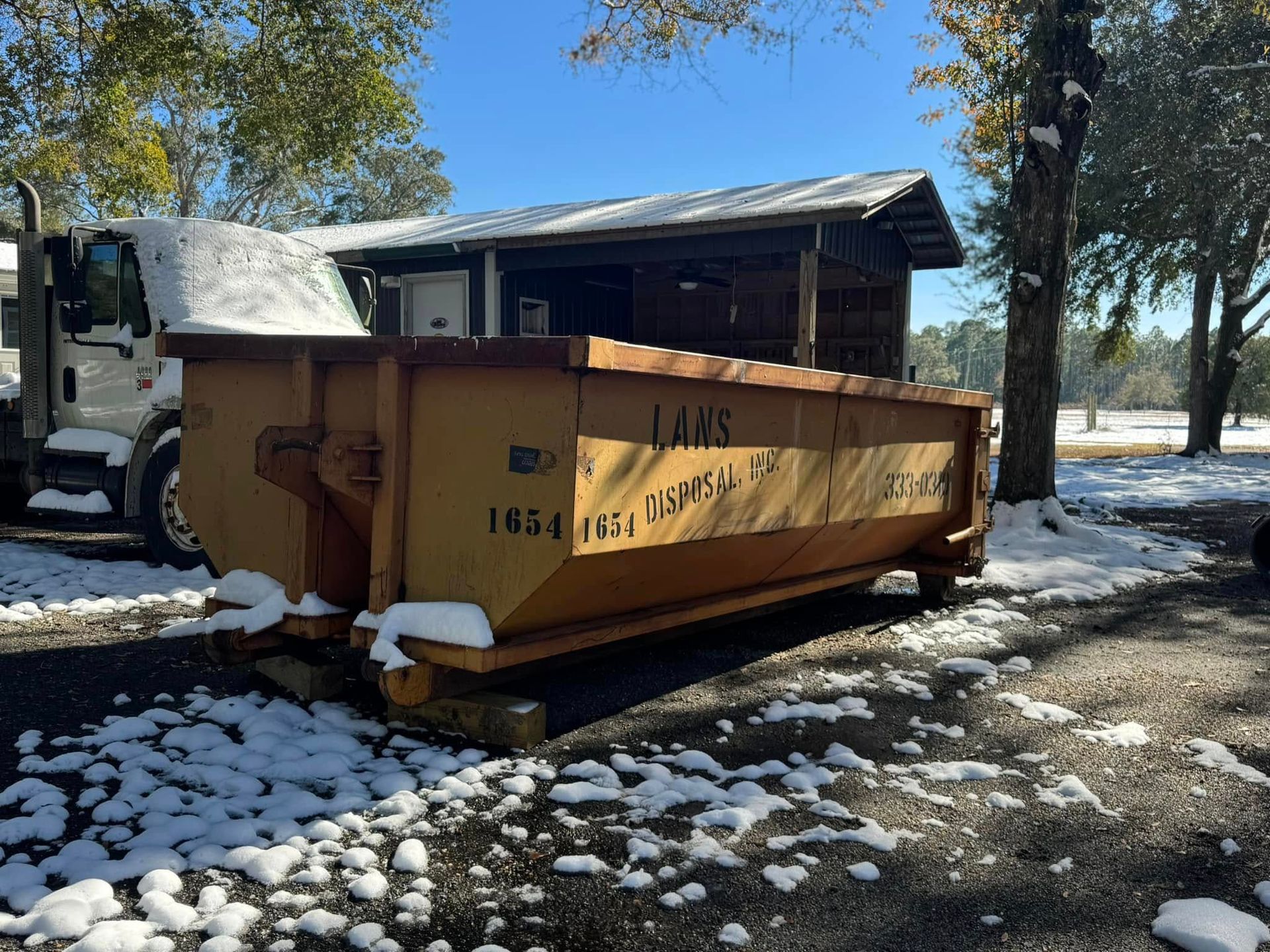 Large orange dumpster beside a truck in a snowy yard under trees