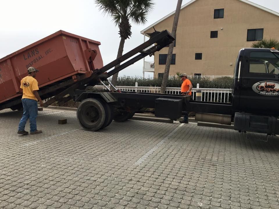 Tow truck lifting a red dumpster in a paved lot, with two workers nearby and a building in the background