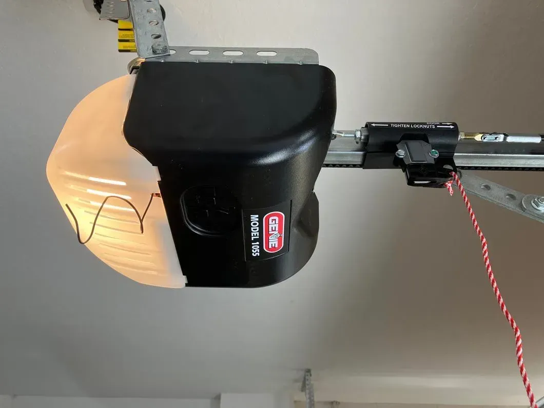 Black and white garage door opener mounted on the ceiling with a light fixture and red wire.