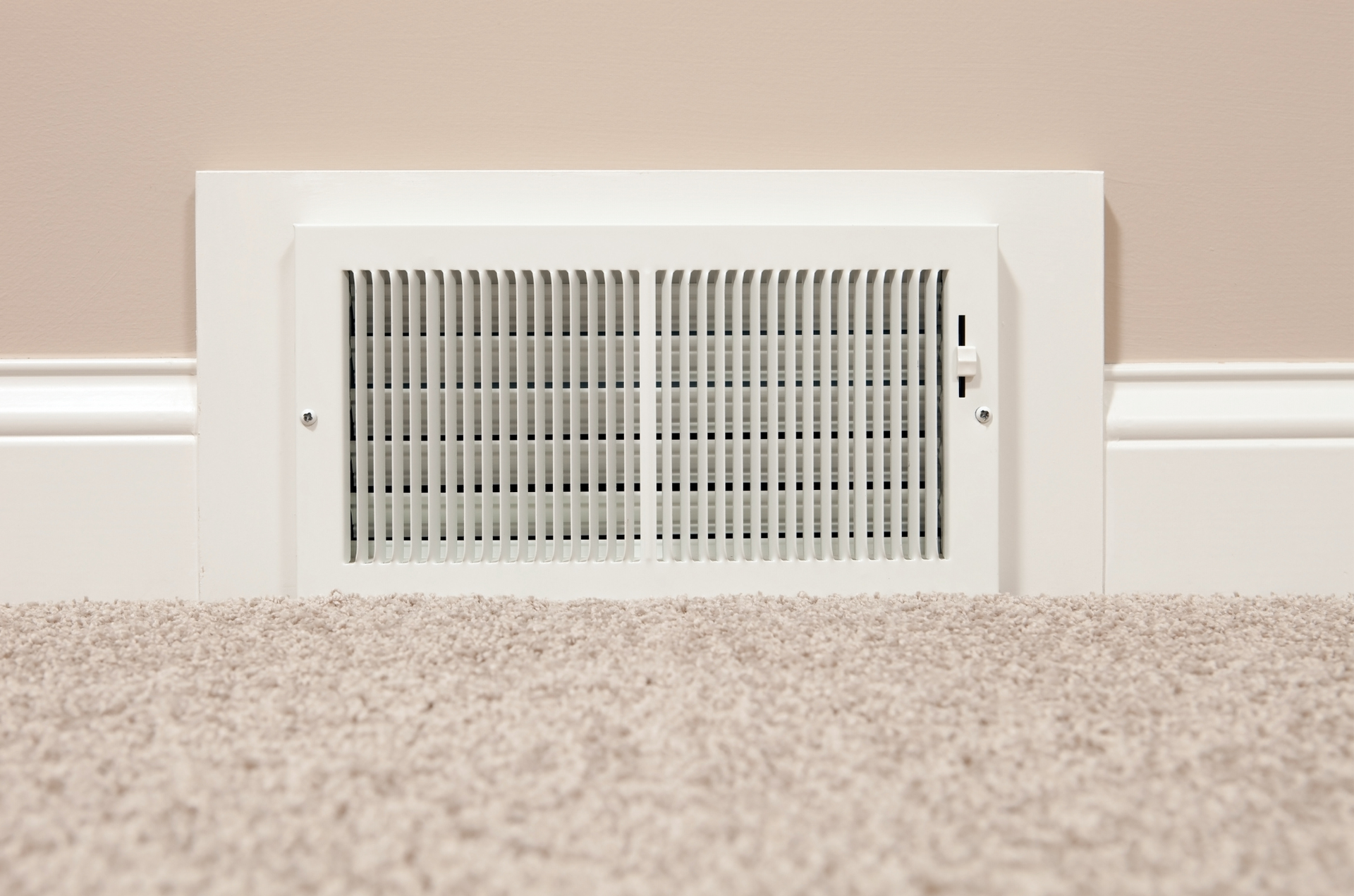 White air vent in a beige wall, installed at the base of the wall with a beige carpet in the foreground.