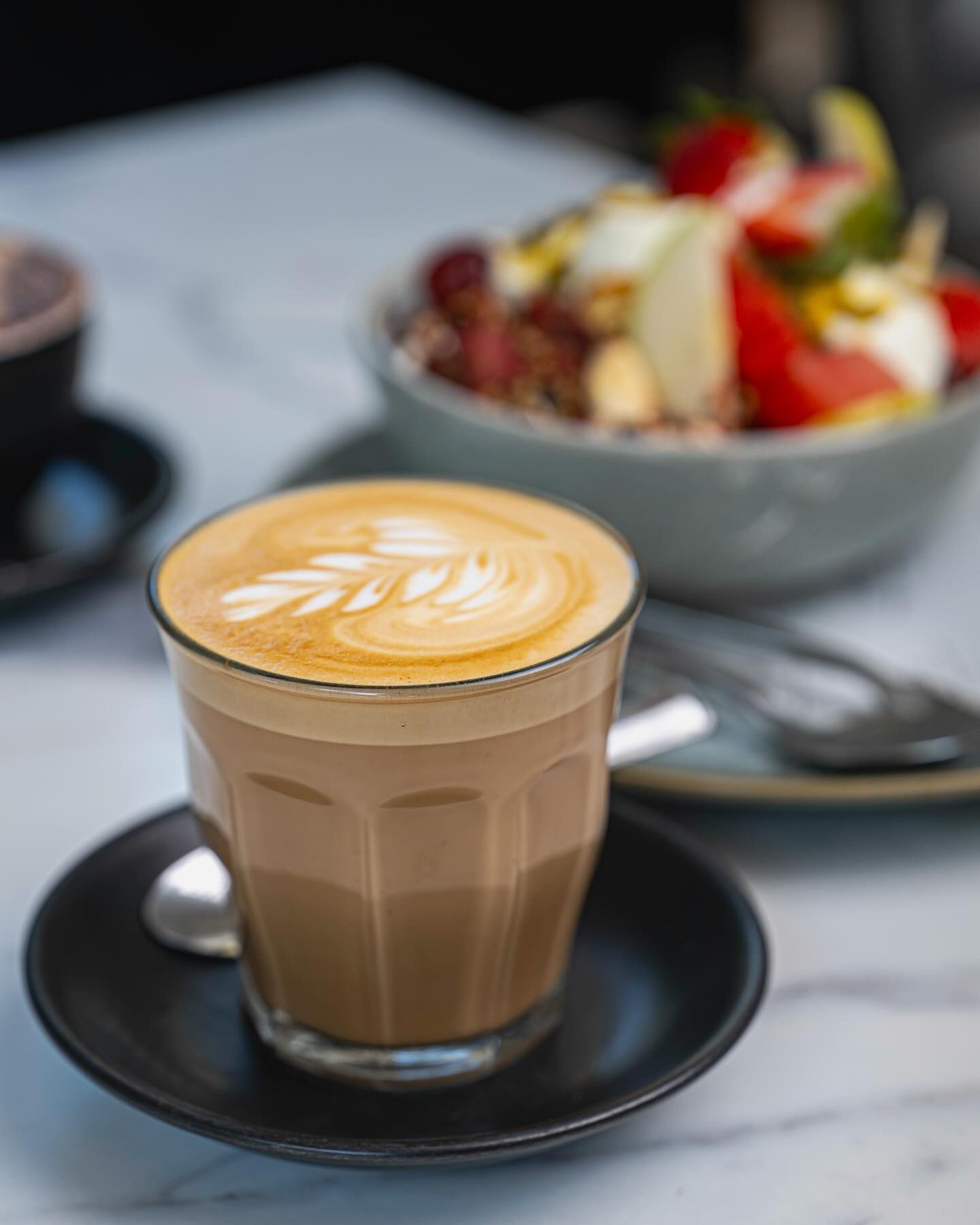 A Cup of Coffee on a Saucer With a Bowl of Fruit in the Background — The Pass Cafe in Byron Bay, NSW