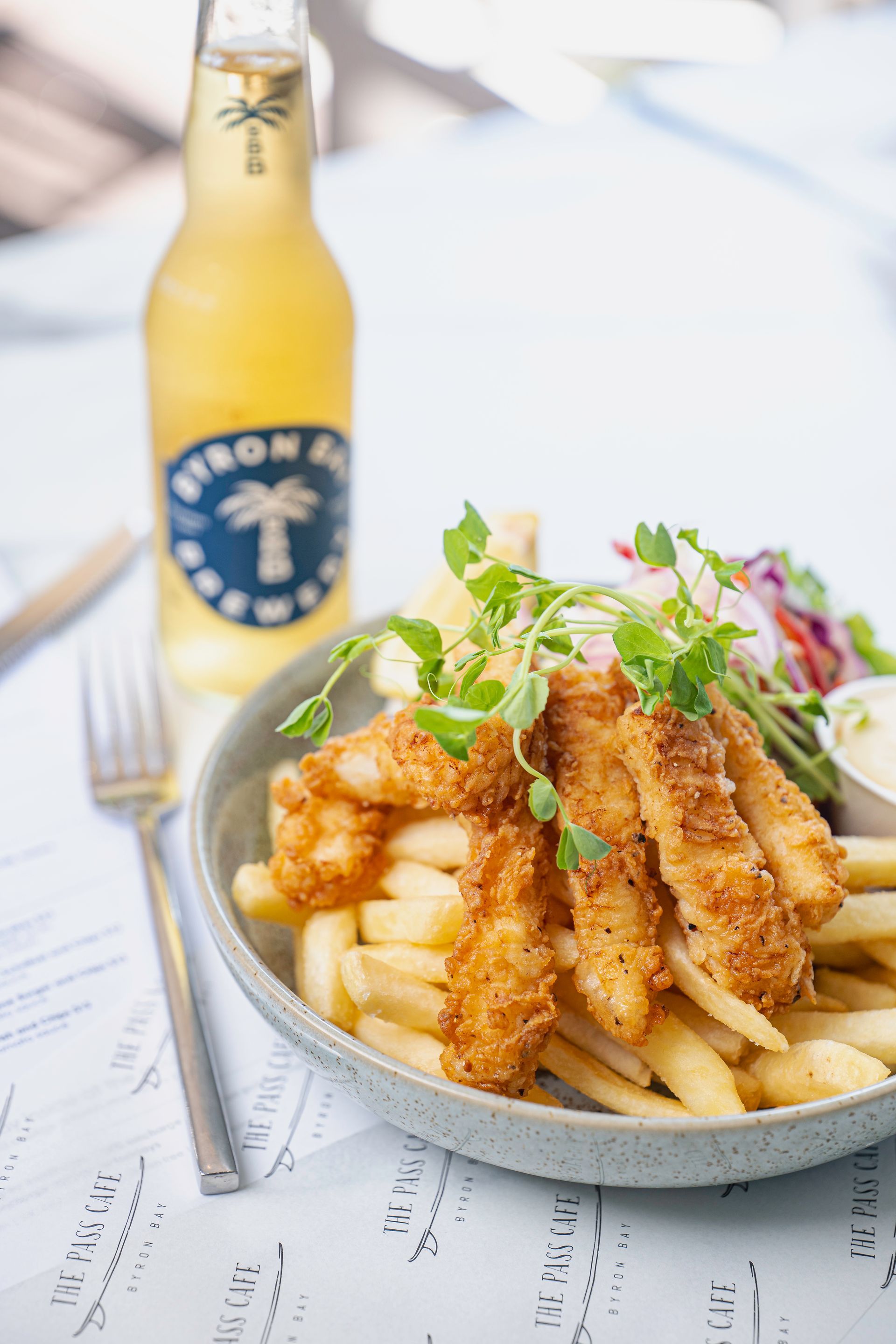 A Bowl of Fried Fish and French Fries Next to a Bottle of Beer on a Table — The Pass Cafe In Byron Bay, NSW
