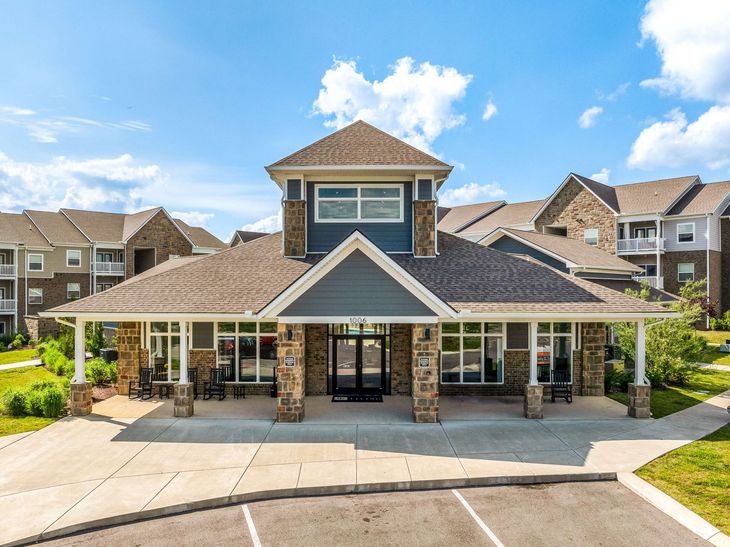 A modern clubhouse exterior with stone pillars and brown roof, next to apartments under a blue sky.