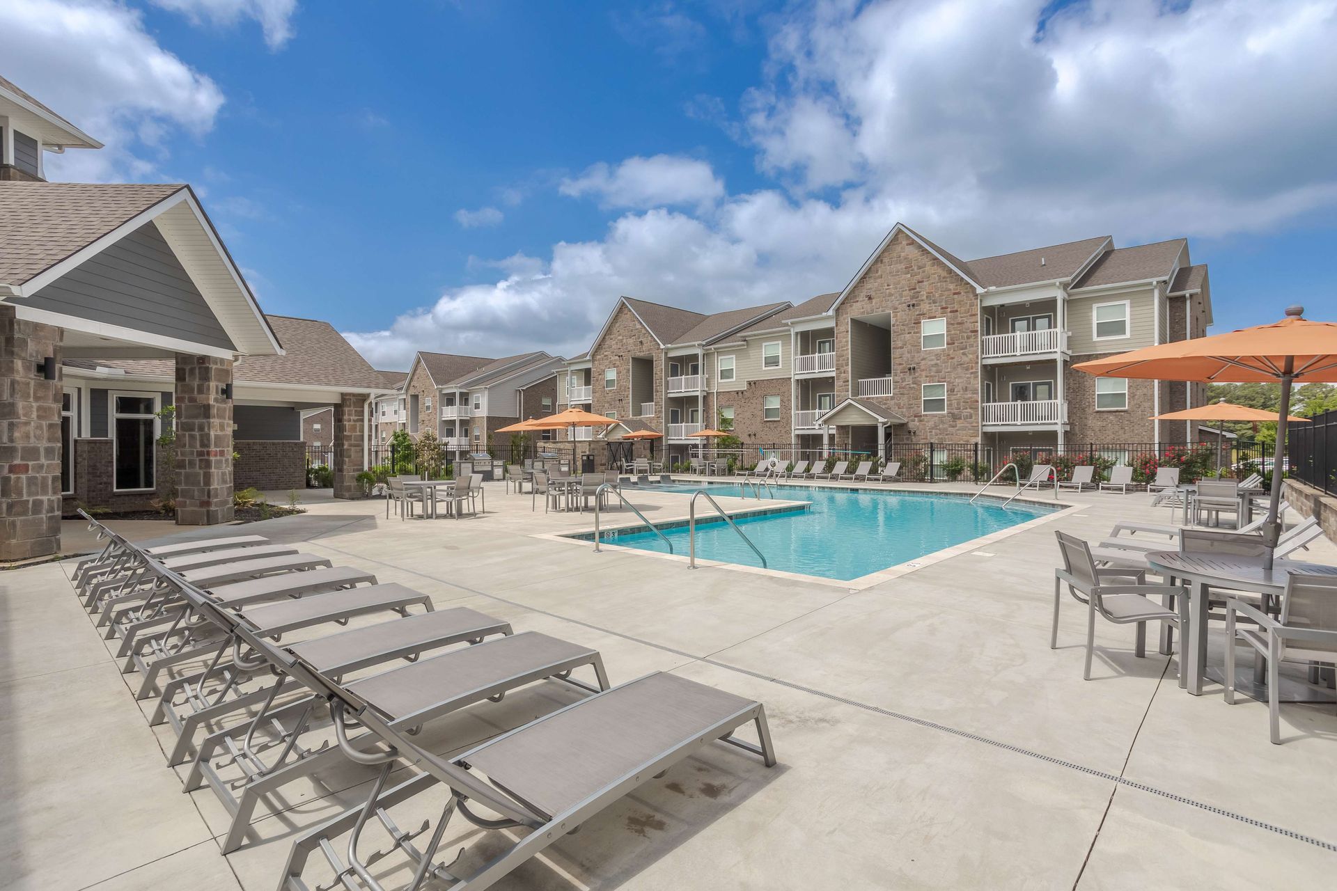 Swimming pool surrounded by lounge chairs, tables, and apartment buildings under a partly cloudy sky.