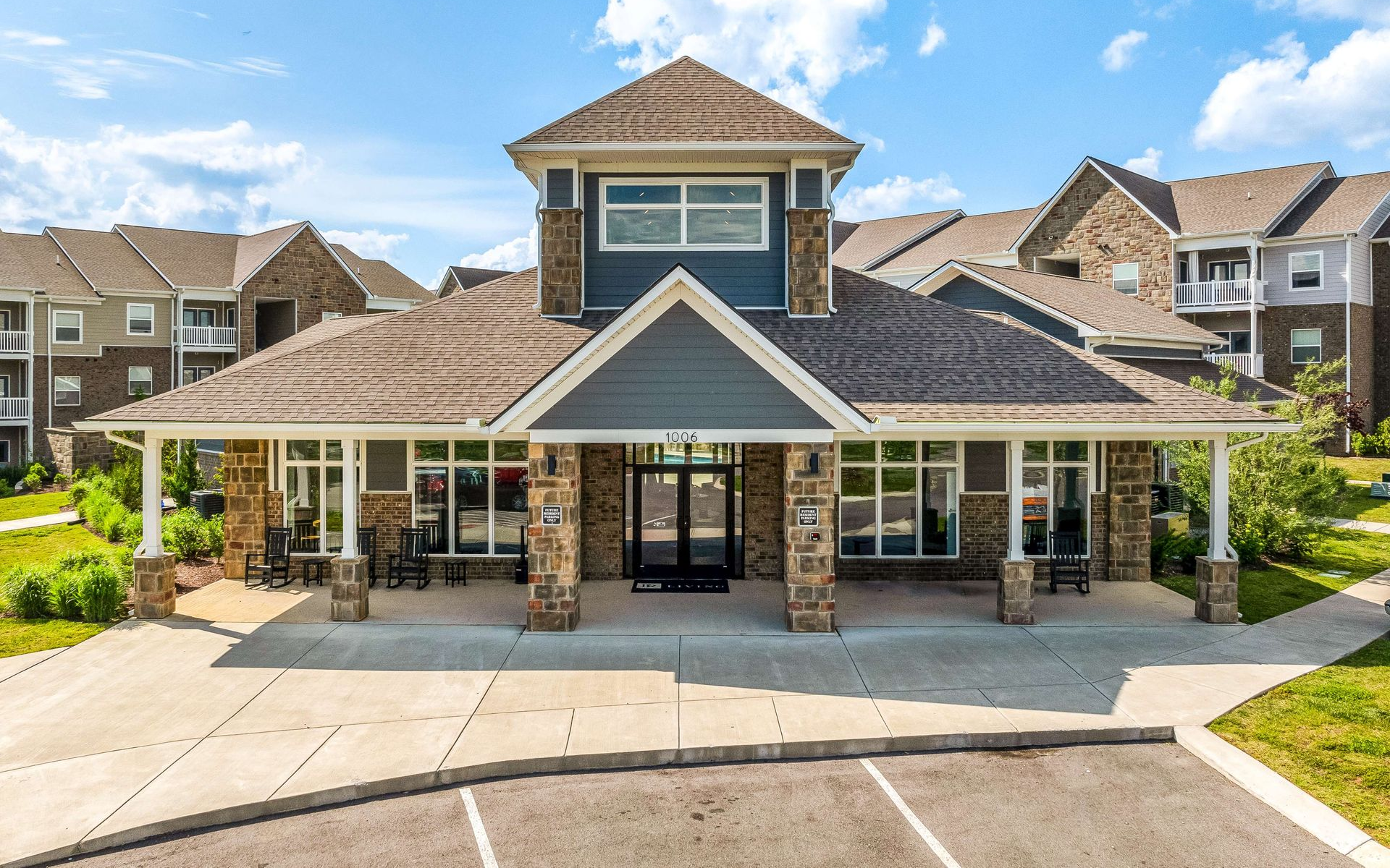 A modern clubhouse exterior with stone pillars and brown roof, next to apartments under a blue sky.