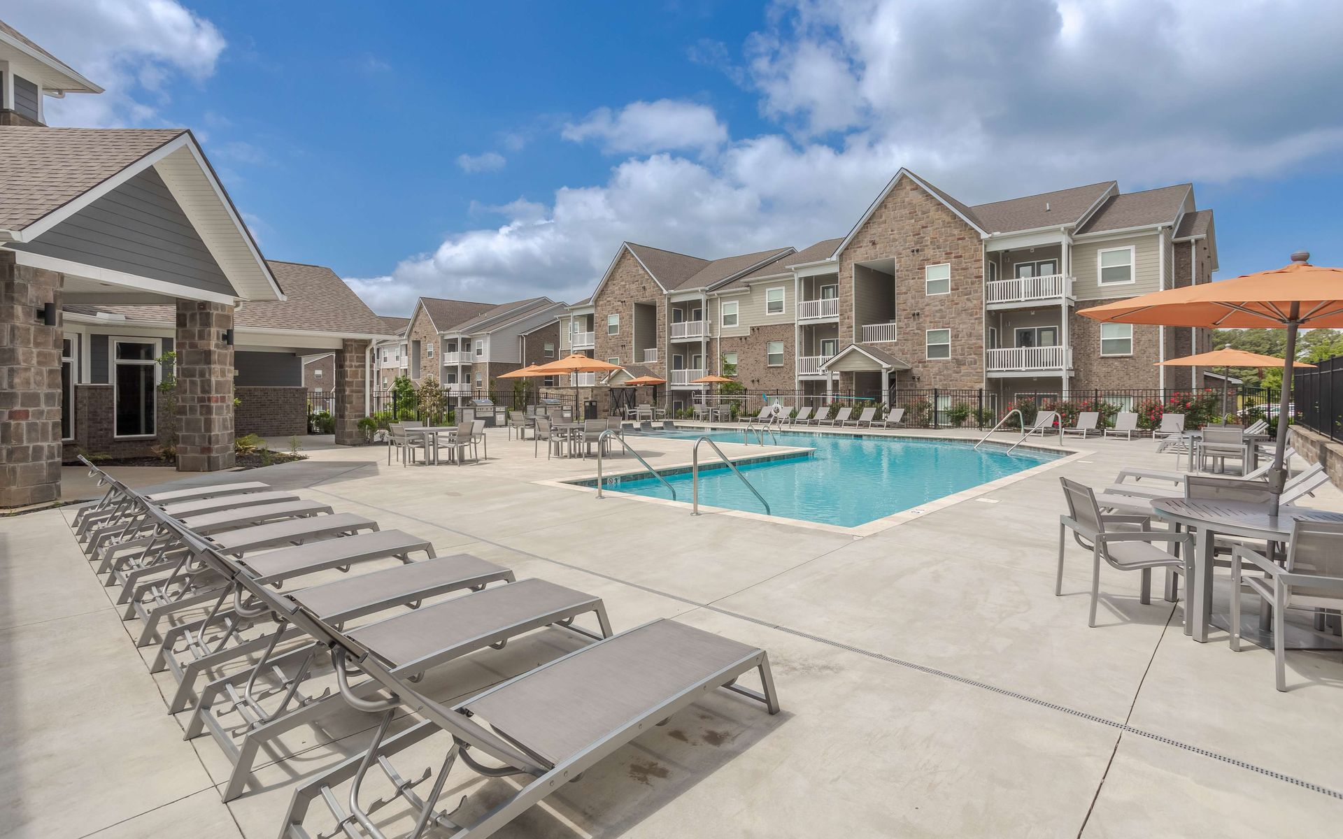 Swimming pool surrounded by lounge chairs, tables, and apartment buildings under a partly cloudy sky.