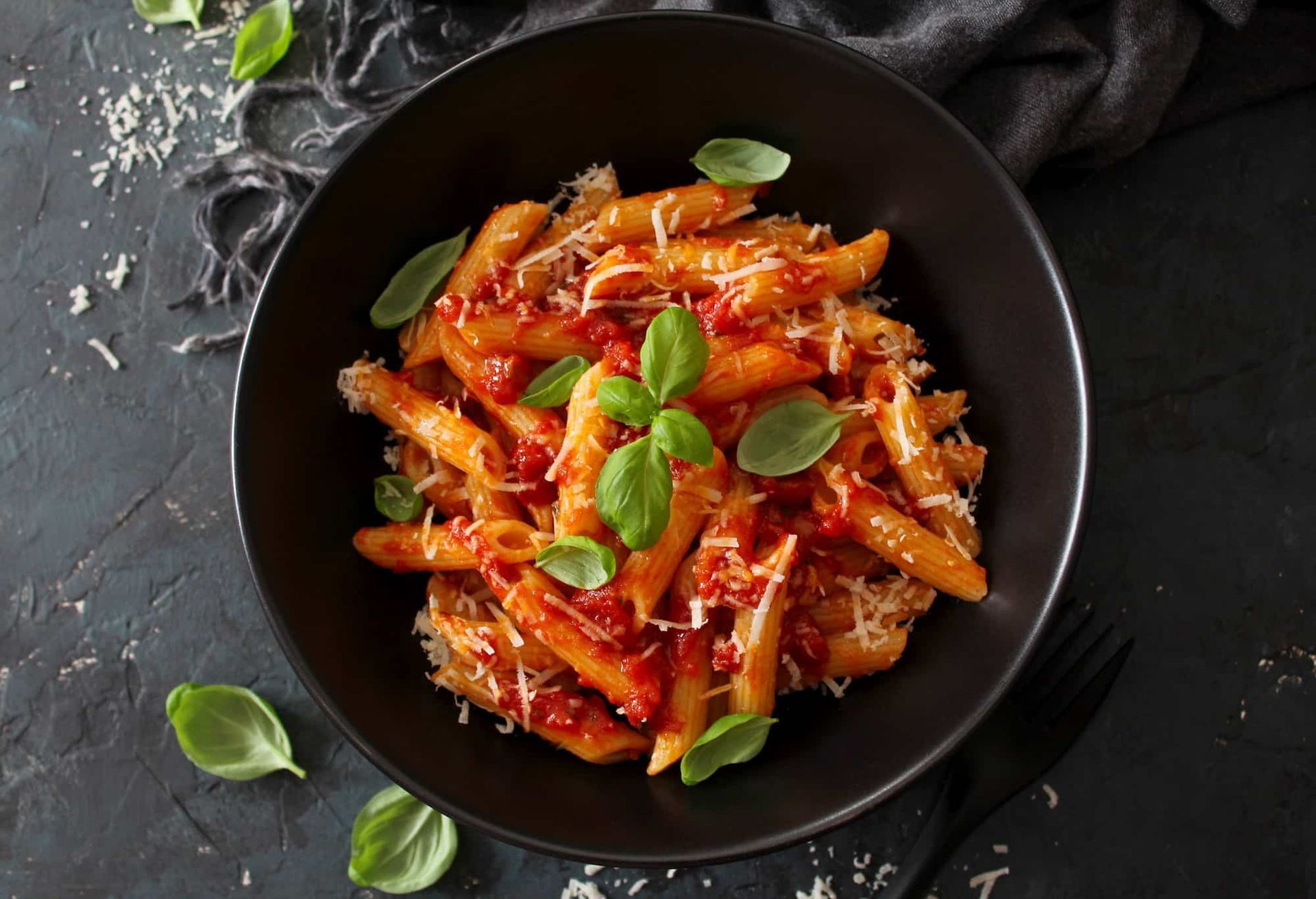 Pasta with tomato sauce, basil, and cheese in a black bowl on a dark surface.