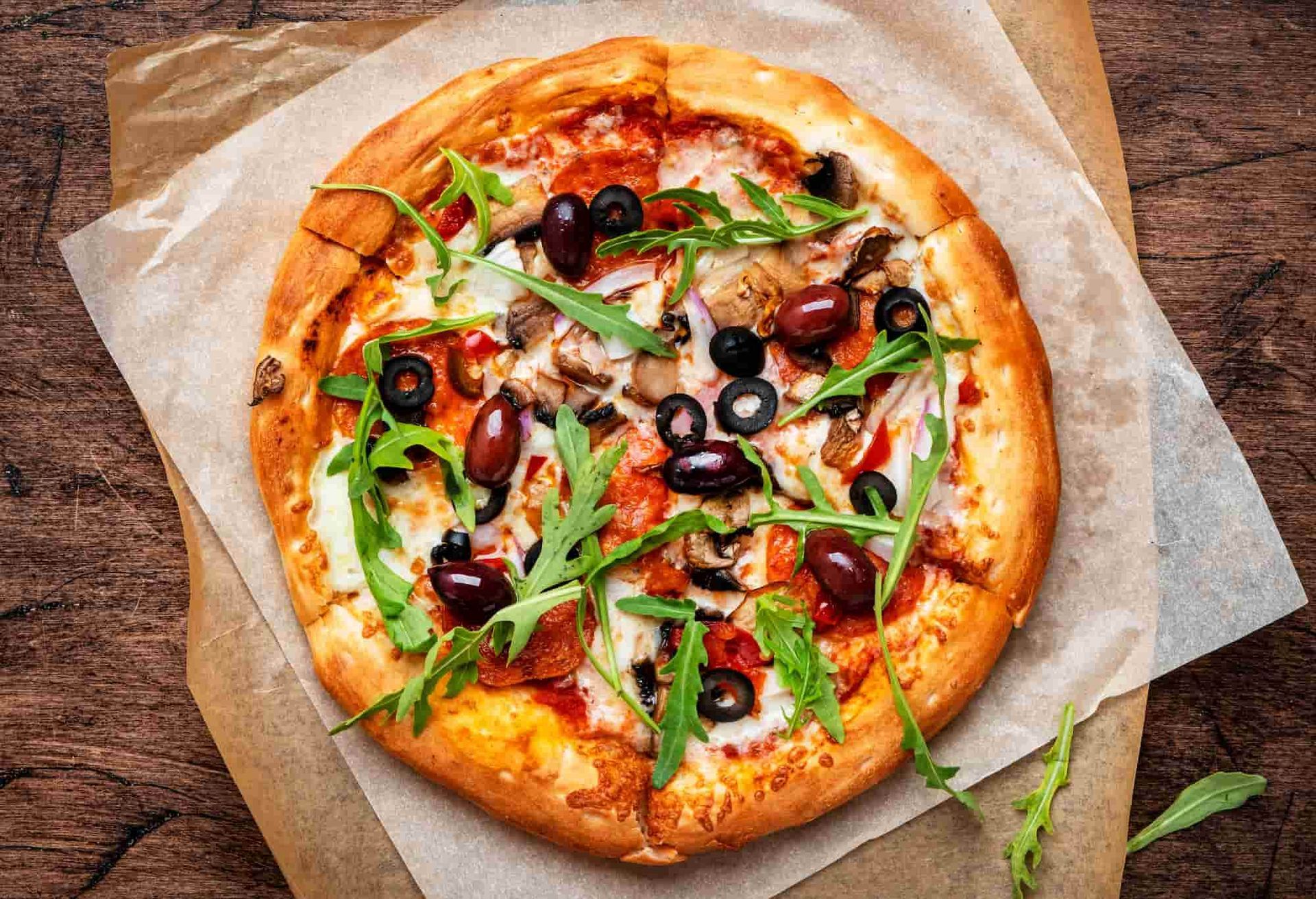 Pizza topped with olives, mushrooms, and arugula on parchment paper atop a wooden surface.