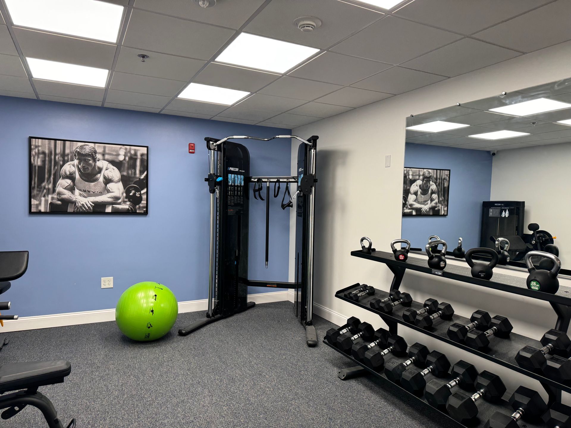 Gym with exercise equipment, including weights, a treadmill, and an exercise ball. Blue walls and bright lighting.