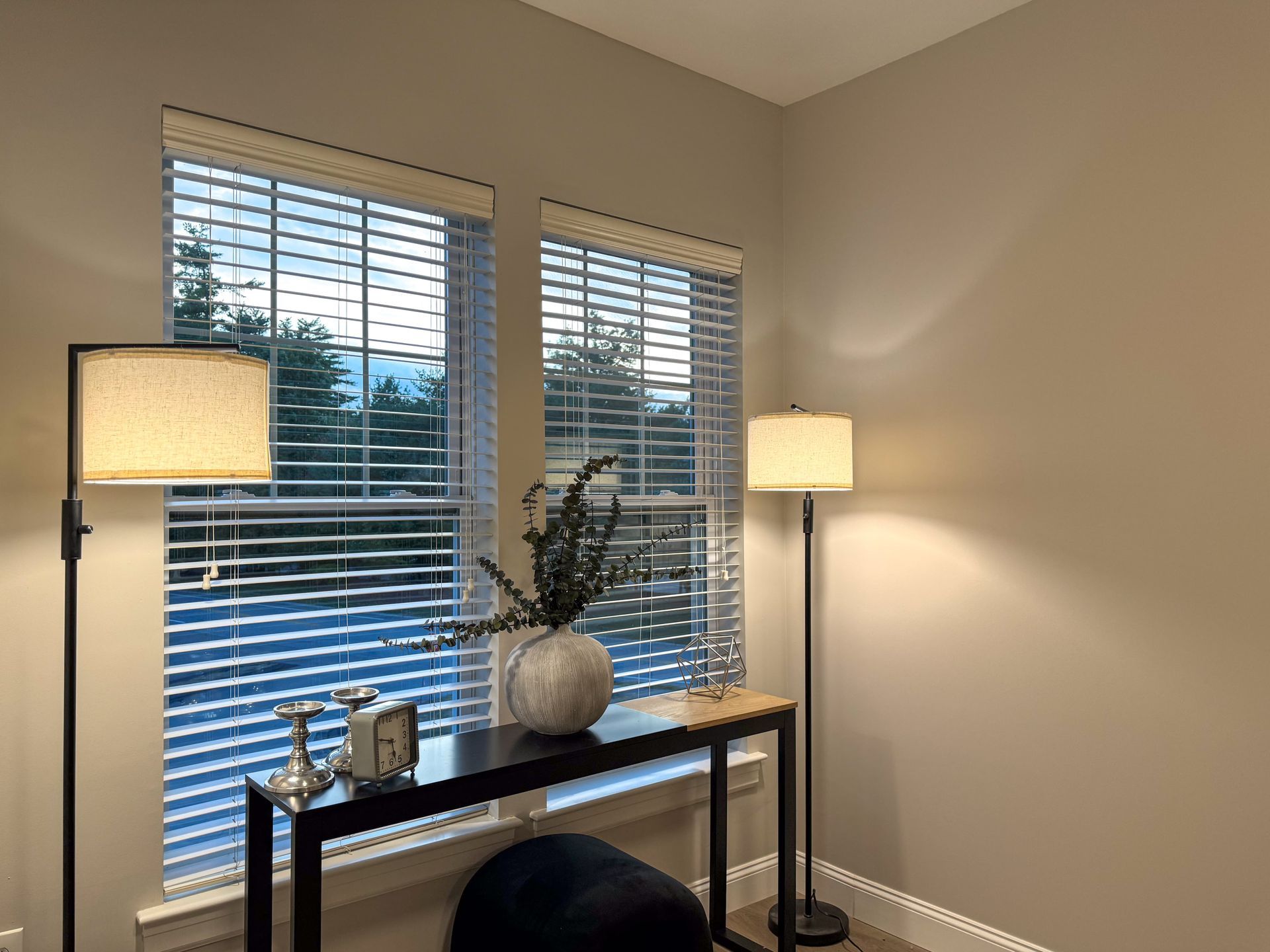 Two windows with blinds, black console table with decor, two lamps on either side.