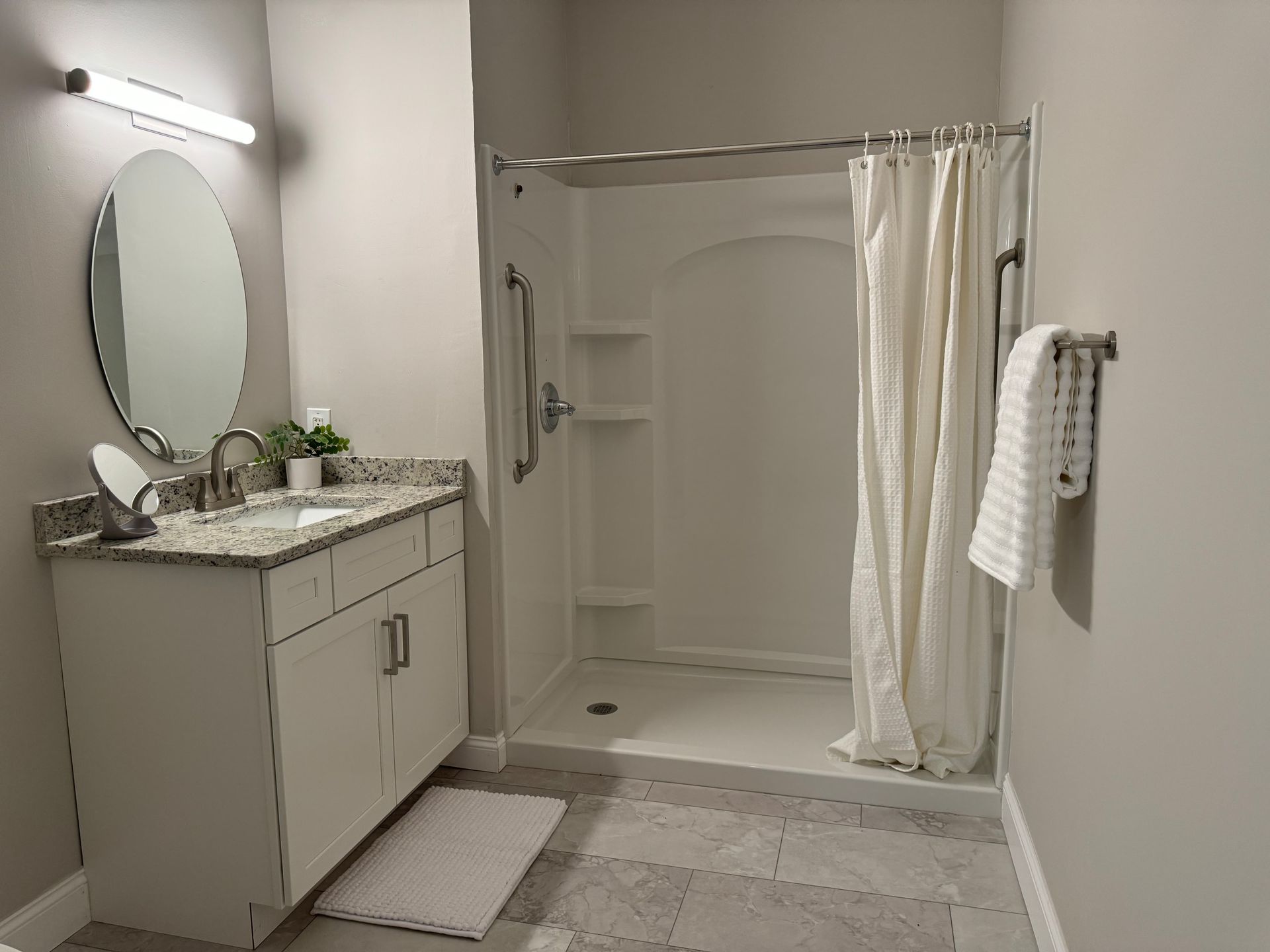 Bathroom with white shower stall, vanity, and neutral-colored walls. A towel hangs on the wall, and a rug sits on the floor.