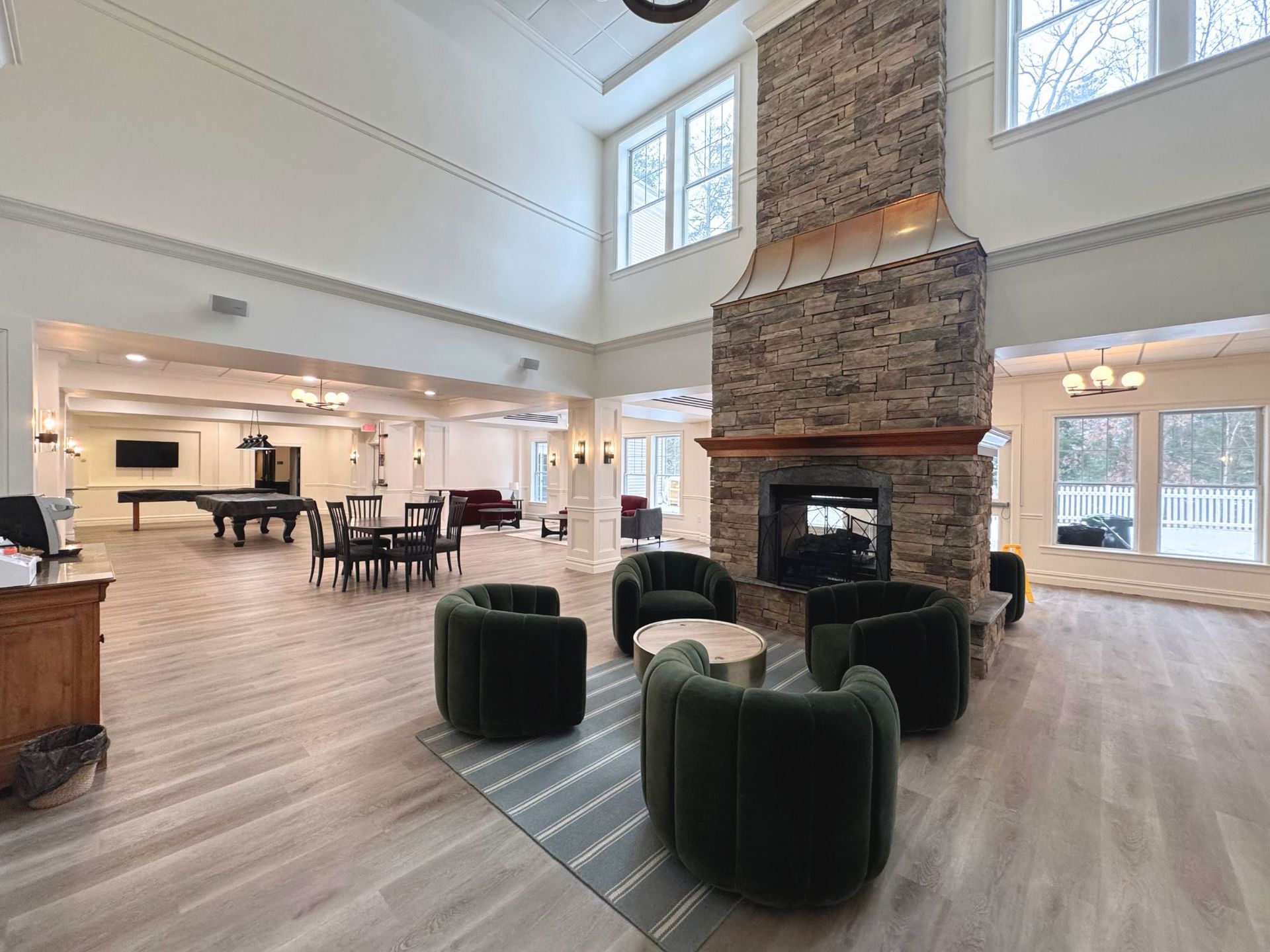 Spacious lounge with fireplace, armchairs, pool table, and tall windows. Light wood floors, white walls.