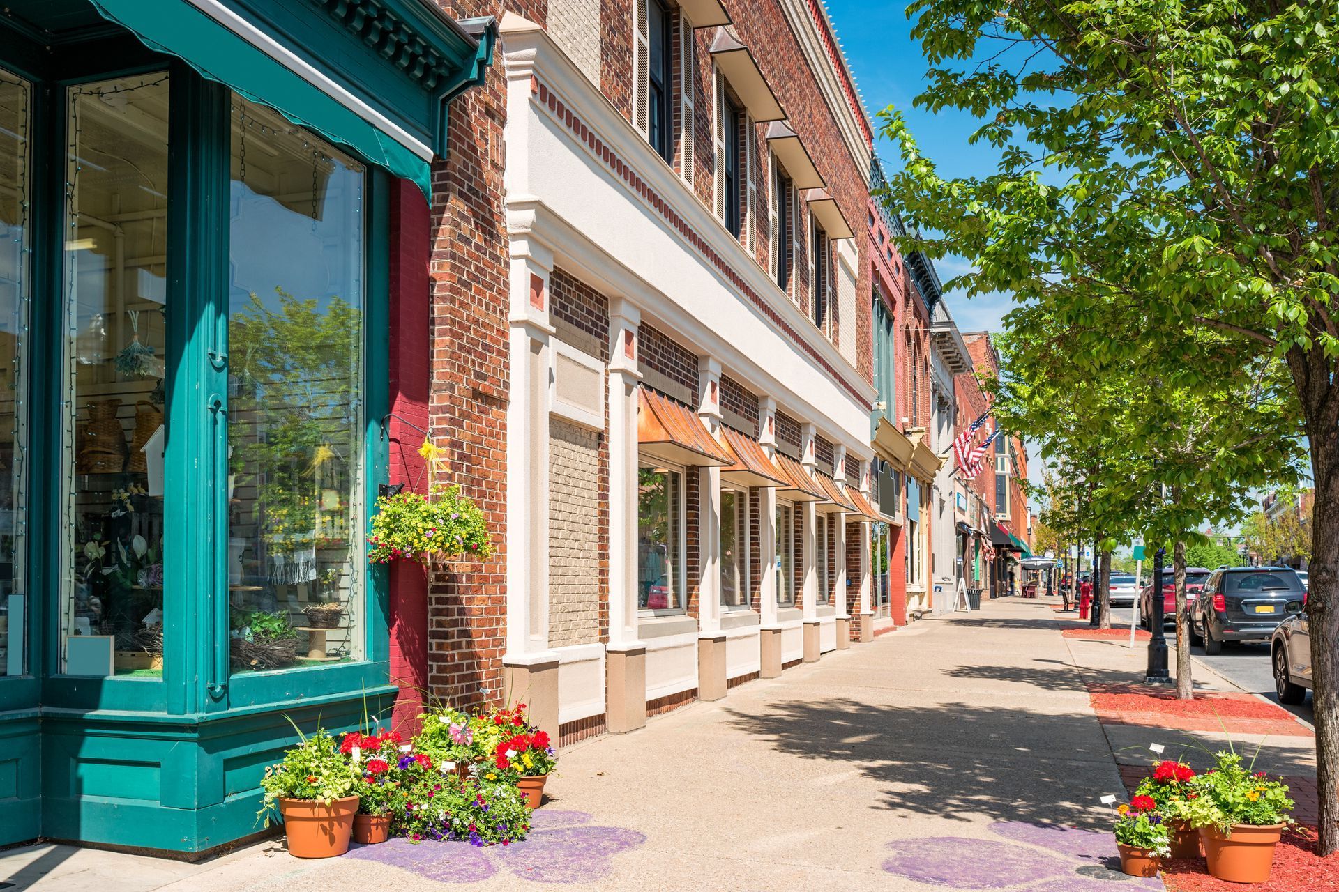 Street with shops, sidewalk, and trees on a sunny day. Teal and brick buildings.