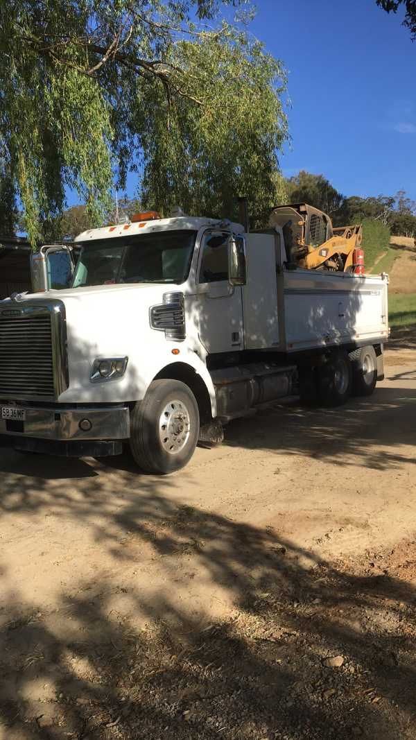 A white dump truck is parked in a dirt lot next to a tree.