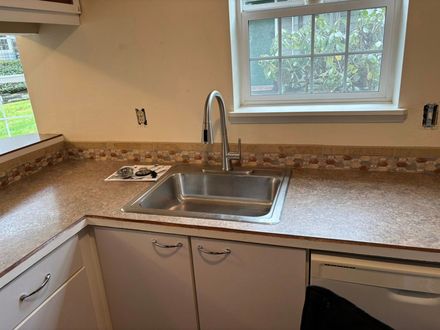 A kitchen sink with a high-arch faucet, set in a light-colored countertop with a tile backsplash beneath a window.